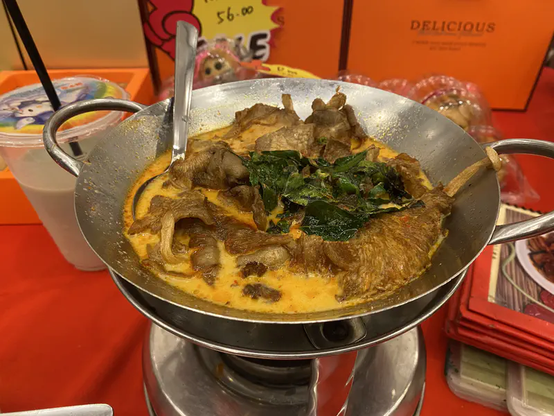 Spicy curry dish served in a metal wok with fried mushrooms and green herbs, accompanied by a drink in a plastic cup with a straw.