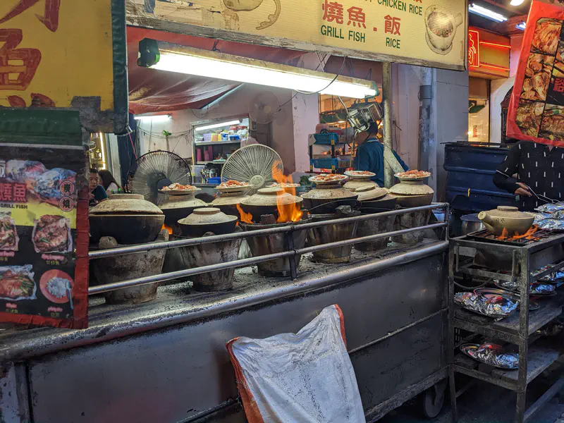 Street food stall with multiple clay pots on open flames, cooking rice and dishes, with signs reading Grill Fish and Chicken Rice above.