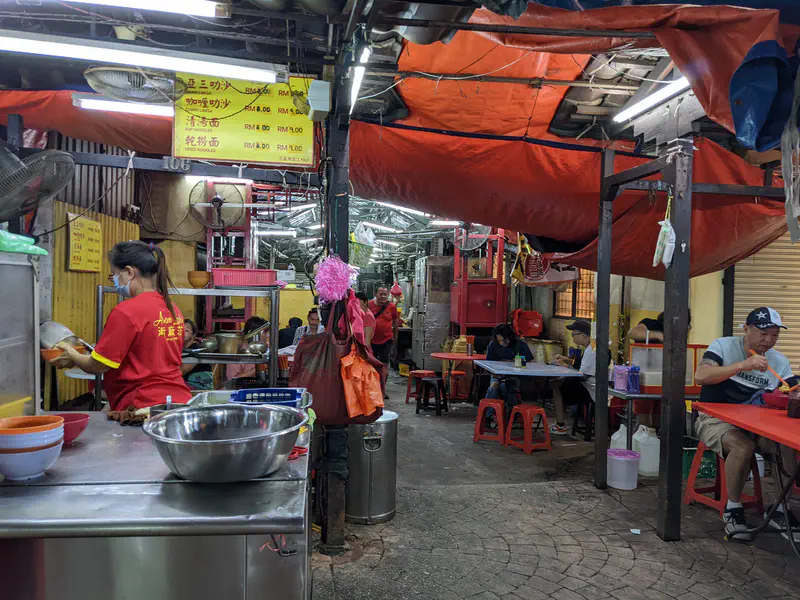 Open-air food stall with red tarps overhead, staff preparing meals, and customers sitting at tables eating noodles.