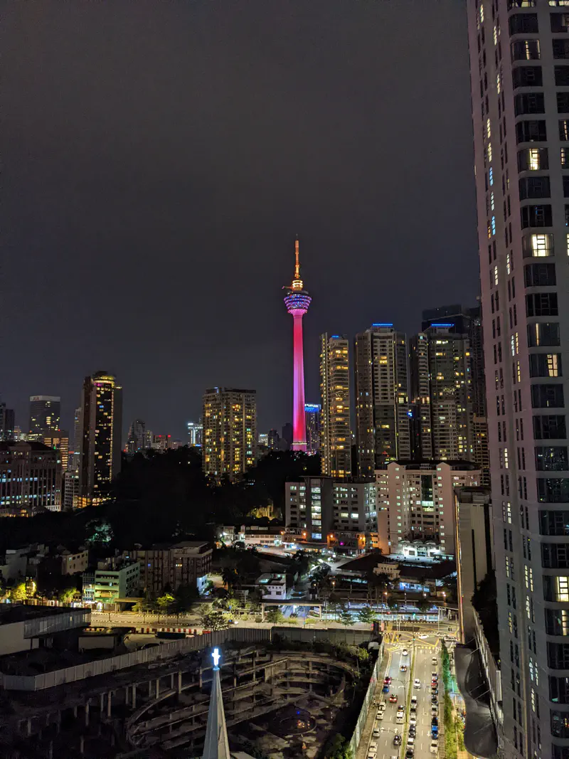 Night cityscape of Kuala Lumpur with the KL Tower illuminated in red and purple, surrounded by tall buildings and busy streets below.