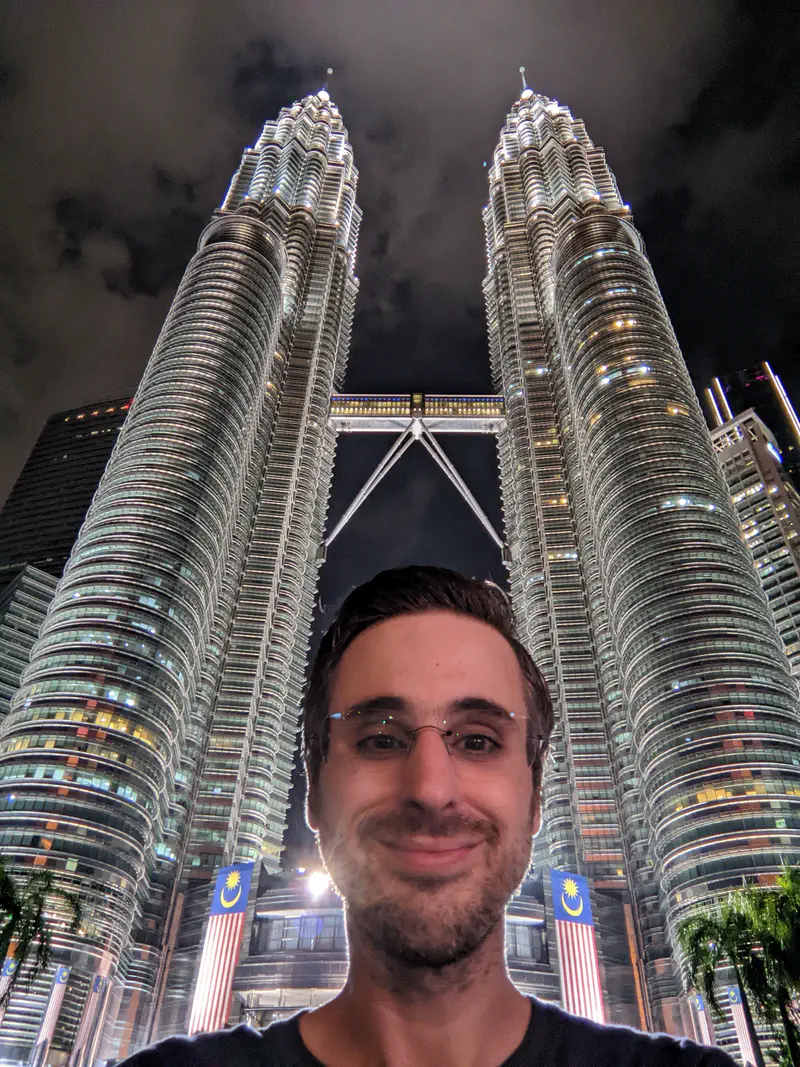 Smiling person taking a selfie at night in front of the illuminated Petronas Twin Towers in Kuala Lumpur, with Malaysian flags visible at the base.