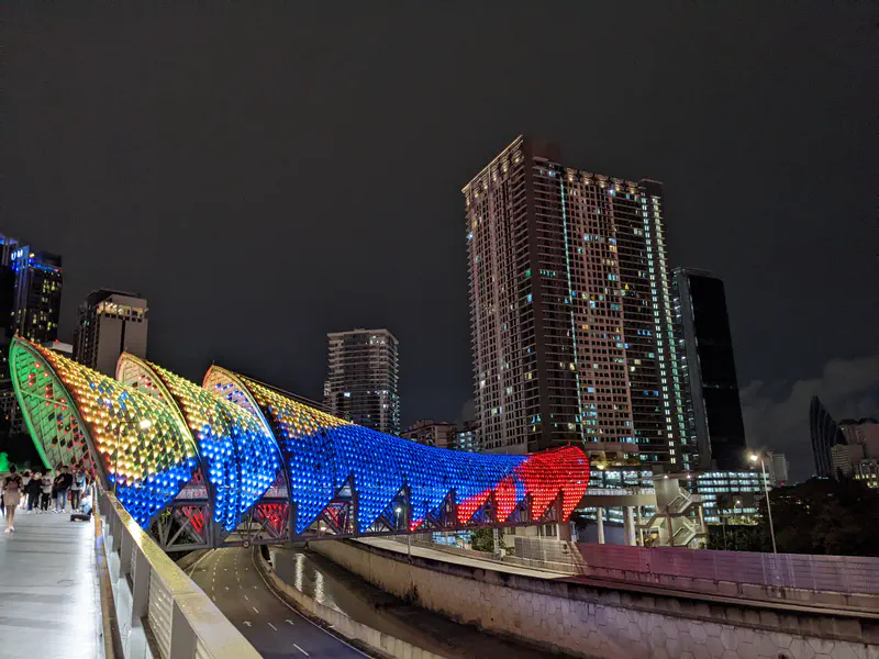Night view of the Saloma Link bridge in Kuala Lumpur, illuminated with colorful LED lights in green, yellow, blue, and red, with tall buildings in the background.
