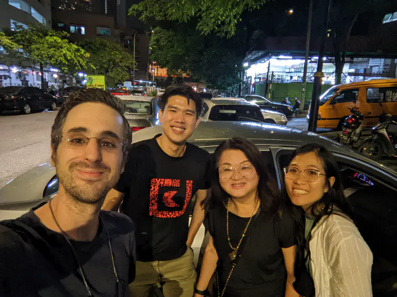 Four friends smiling and posing for a group photo at night in a parking lot with cars and streetlights in the background.