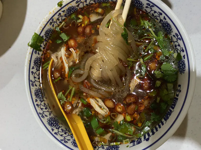 Spicy noodle soup with translucent noodles, shredded chicken, peanuts, chili oil, and fresh herbs in a patterned bowl.