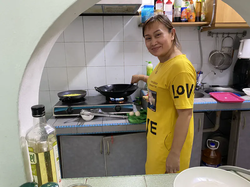 Woman in a yellow shirt cooking at a gas stove in a kitchen, smiling at the camera.
