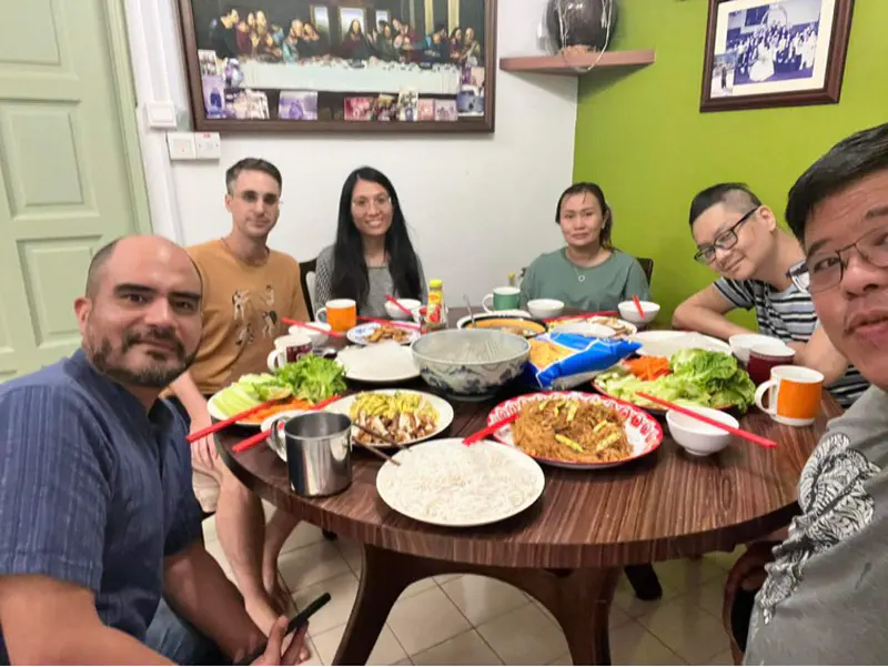 Six people sitting around a table sharing a meal with noodles, vegetables, and drinks.