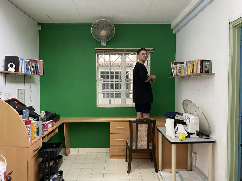 Man standing on a chair arranging books on a shelf in a room with a green wall and desk.