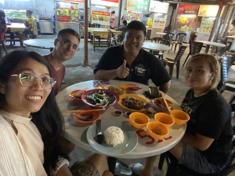 Four people sitting at a table with rice, noodles, and vegetables at an outdoor food court.