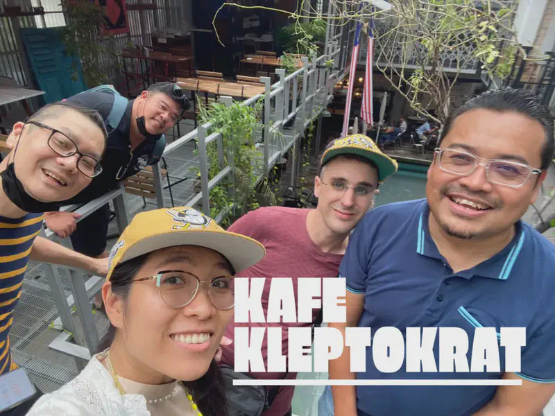 Group selfie of five people smiling at Kafe Kleptokrat with outdoor seating and a pool in the background.