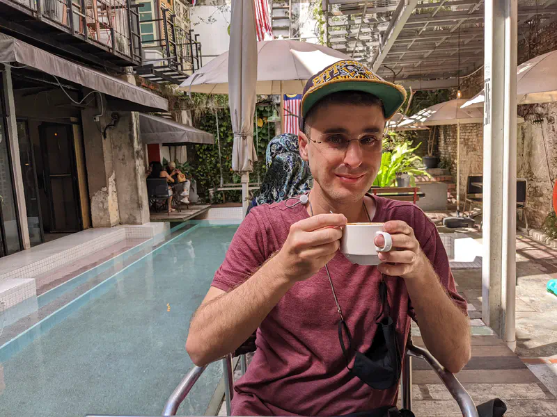 Man wearing a cap holding a cup of coffee at an outdoor café by a pool.
