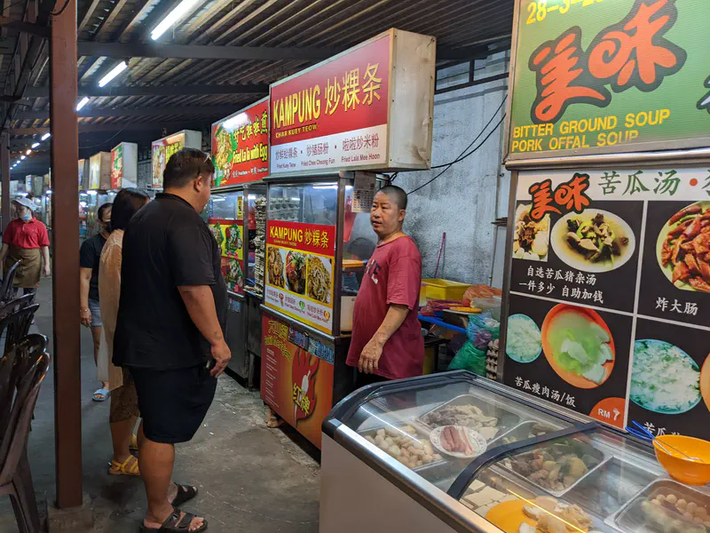 Man ordering food at a hawker stall with bright signs and food displays in a night market.