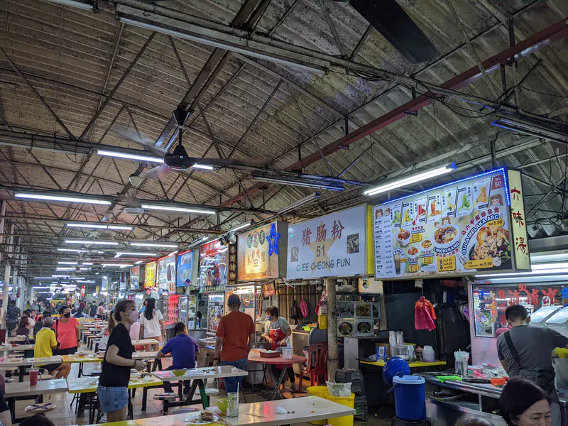 Busy hawker center with food stalls, signs, and people seated at tables under a high ceiling.