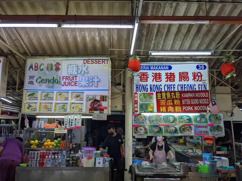 Hawker stall signs for cendol, fruit juice, and Hong Kong chee cheong fun with vendors preparing food.