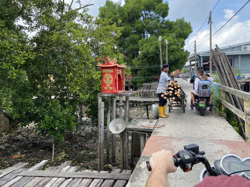 Narrow village pathway with a small red shrine, a man pushing a cart of logs, and people riding scooters.
