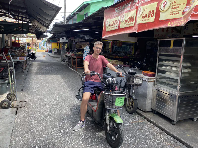 Man in a red shirt and blue shorts sitting on a motorbike in a market street with stalls on both sides.
