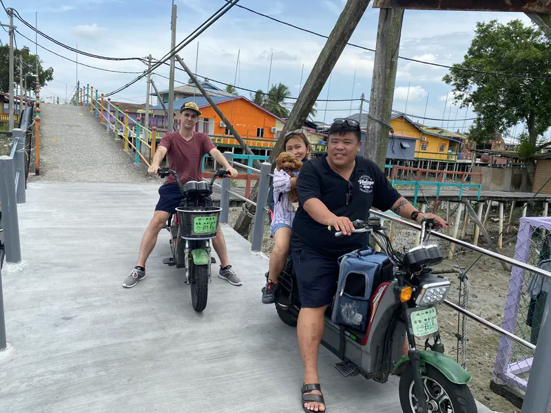 Two people riding motorbikes on a bridge in a fishing village, one holding a small brown poodle, with colorful stilt houses in the background.