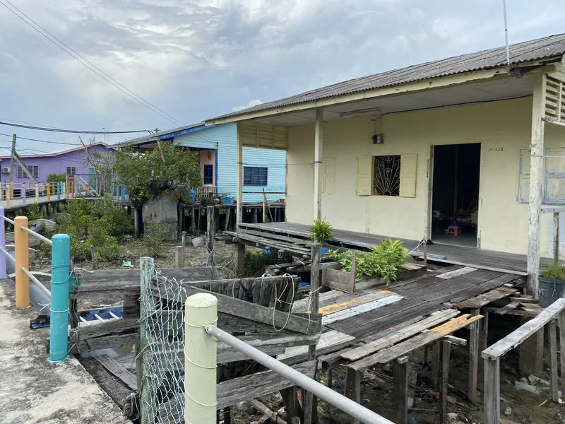 Wooden stilt house with open doorway and plants on a weathered walkway, surrounded by other colorful houses in the background.