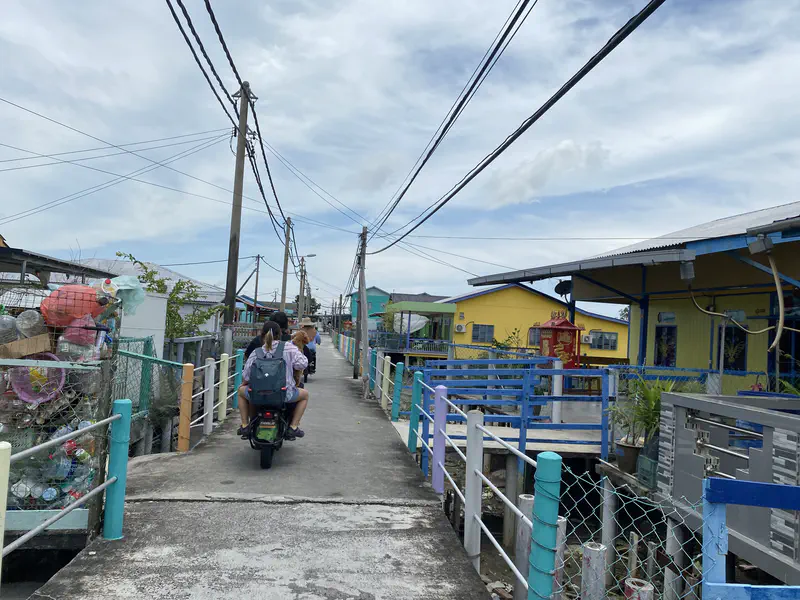 Narrow concrete walkway in a stilt village with two people riding a motorbike, colorful houses and utility poles lining the path.