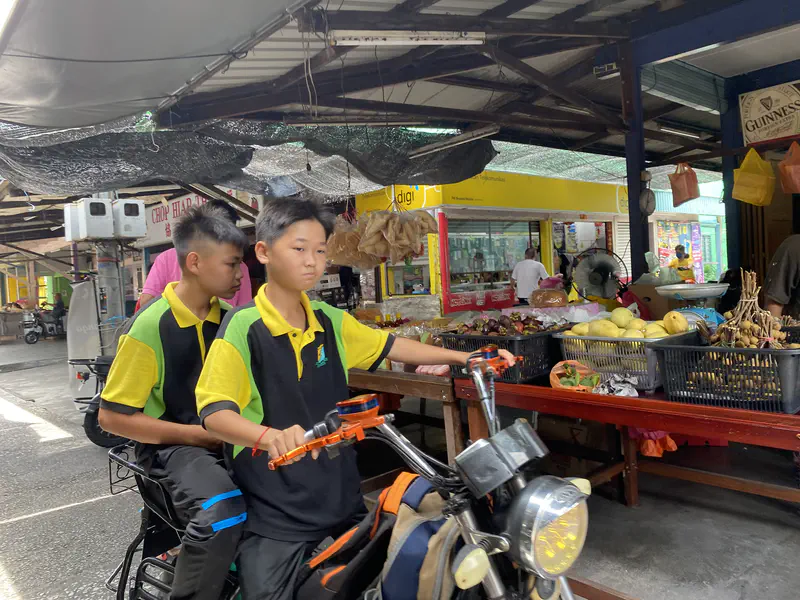 Two schoolboys in black-and-yellow uniforms riding an electric scooter through a covered market beside fruit stalls.