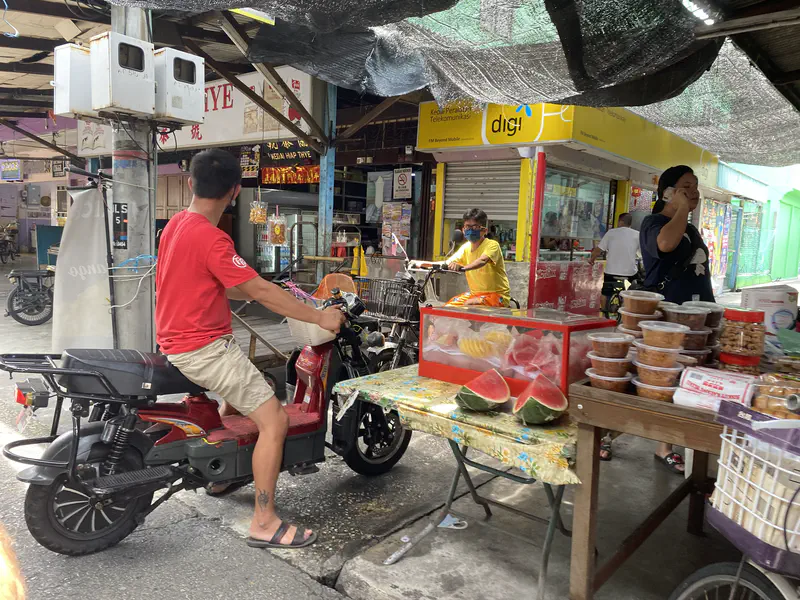 Local market scene with a man on a motorbike, fruit stalls displaying watermelon and packaged goods, a woman on the phone, and shoppers moving through the narrow aisle.