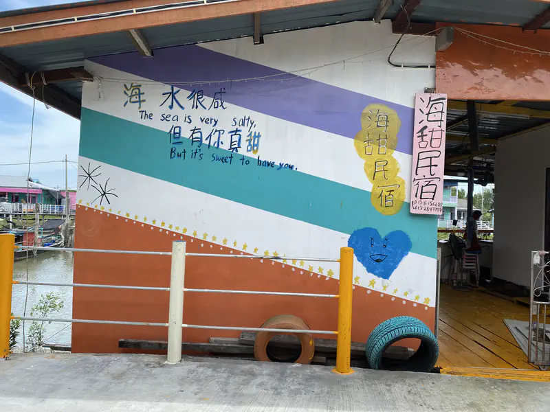 Wall mural in a stilt village with text reading 'The sea is very salty, but it's sweet to have you,' decorated with stars and hearts.