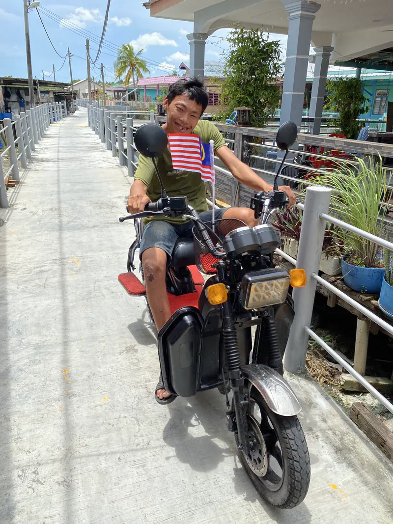 A smiling man riding a motorbike decorated with a Malaysian flag on a narrow concrete path beside houses.