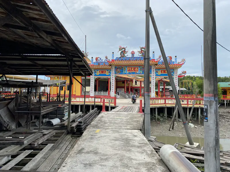 A colorful Chinese temple with ornate dragon decorations on the roof, viewed from a concrete walkway over stilts with wooden planks and poles in the foreground.