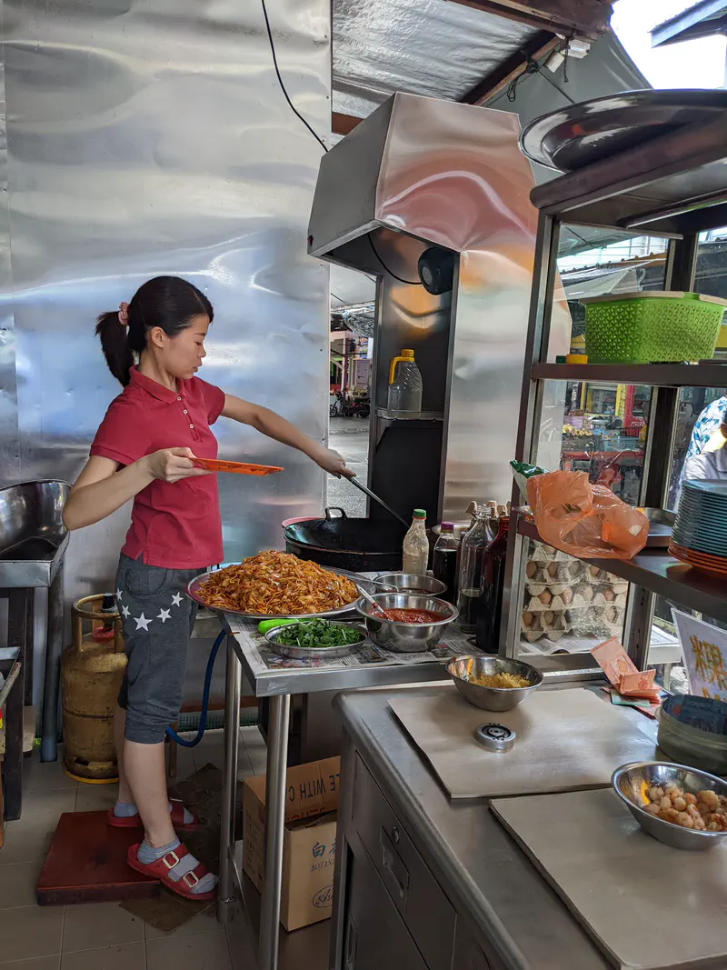 Street-food vendor stir-frying noodles in a wok at a stainless-steel stall, ingredients laid out in bowls.
