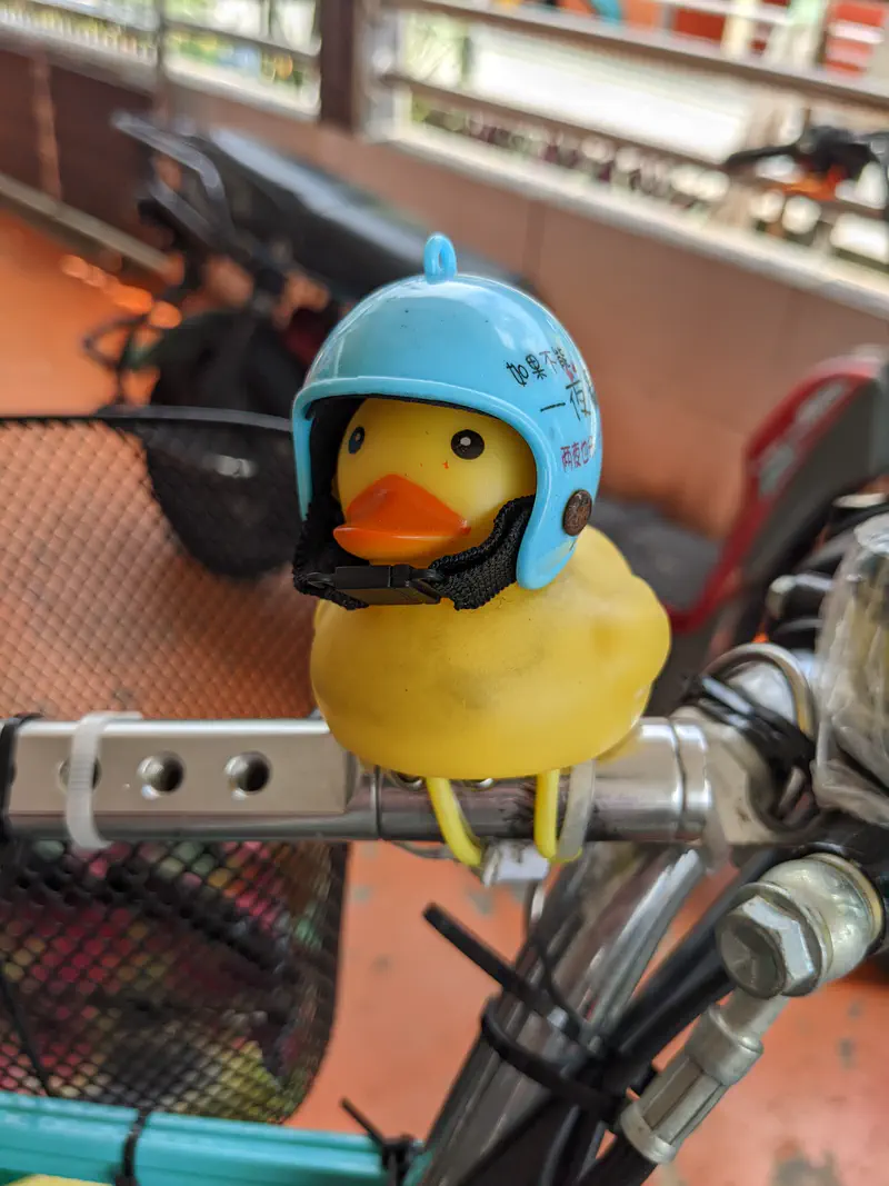 Close-up of a yellow rubber duck wearing a blue helmet, strapped onto the handlebar of a bicycle or scooter.