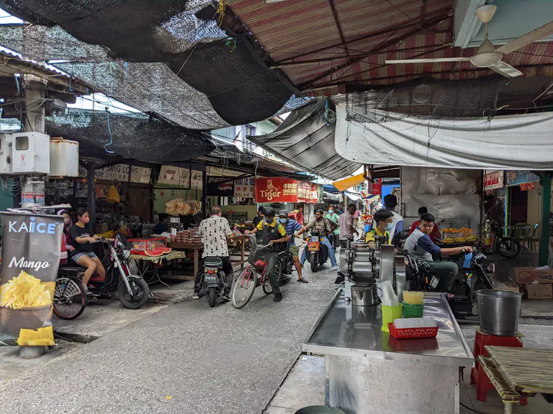 Busy local market with motorbikes weaving through narrow aisles, fruit and food stalls on both sides, and a vendor preparing a large plate of noodles at a cooking station.