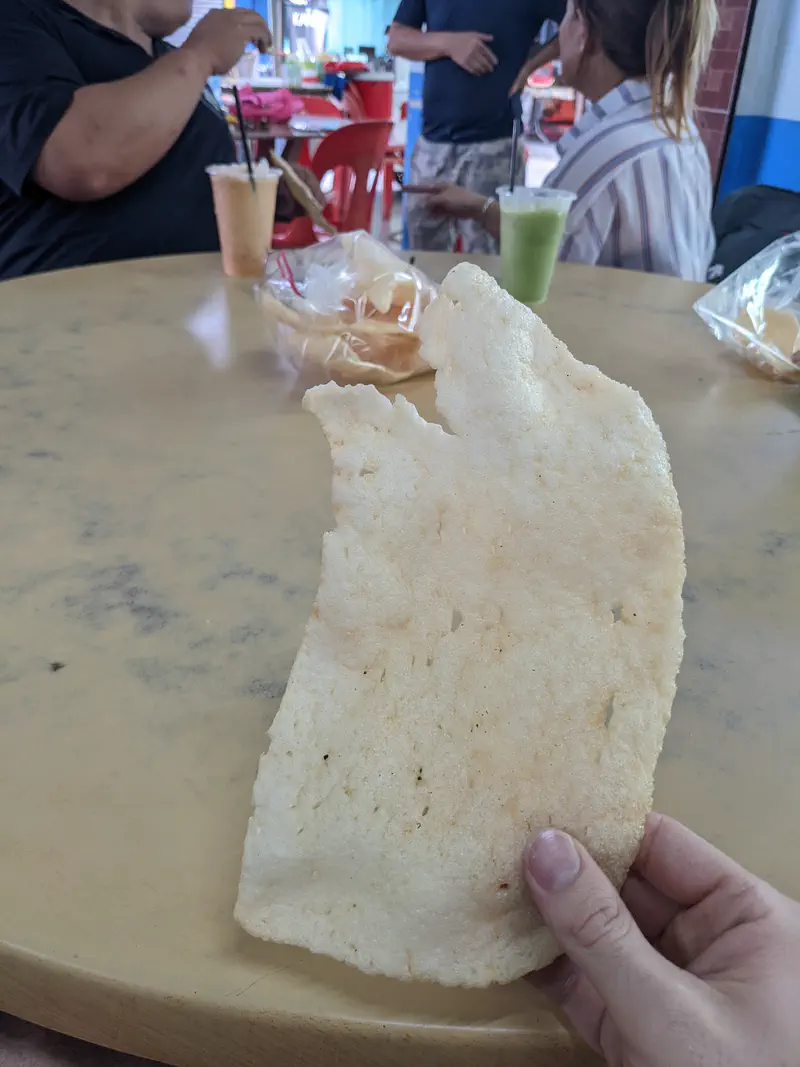 A person holding a large crispy cracker at a round table with drinks and snacks, while others sit and chat in the background.