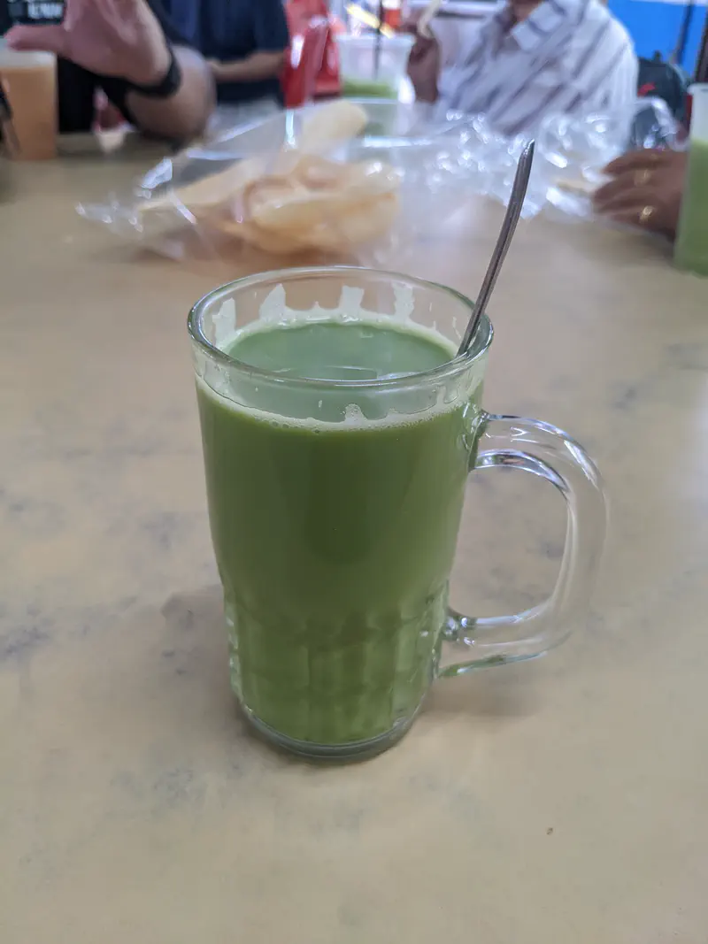 A glass mug filled with freshly pressed green sugarcane juice on a table with people and snacks in the background.