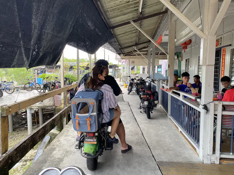 Two people riding a scooter along a narrow stilt walkway past houses where locals sit and chat.