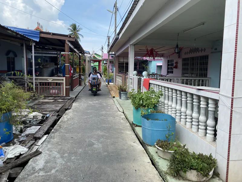A narrow concrete walkway between stilt houses, with a person riding a scooter carrying a passenger wearing a blue backpack.