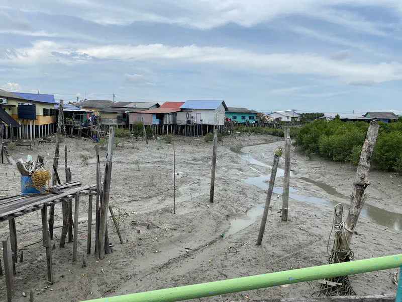 Stilt houses with colorful roofs line the edge of a muddy riverbed at low tide, with small streams of water running through the mud.