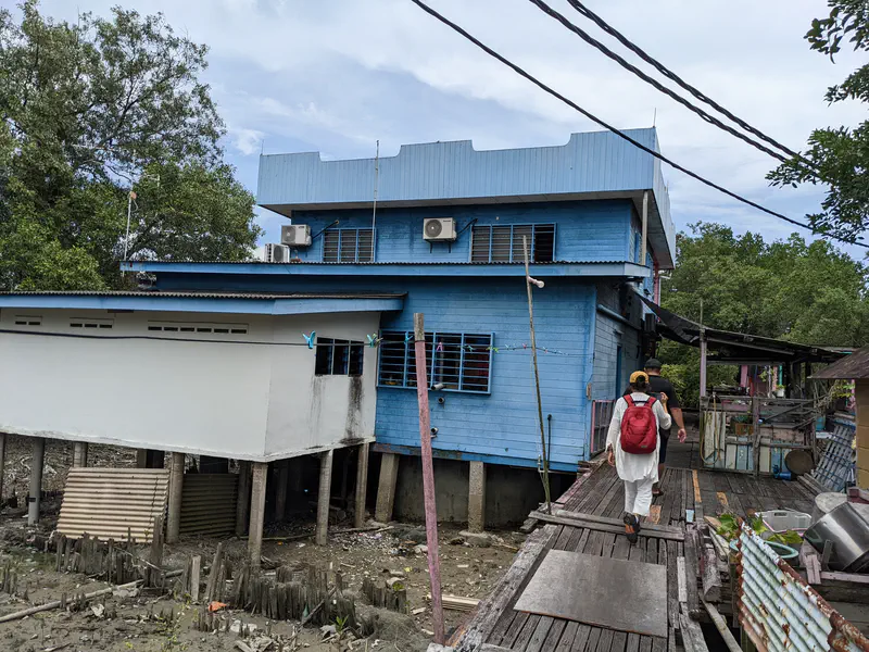 People walking along a wooden walkway towards a large blue stilt house with air conditioners mounted on the walls.