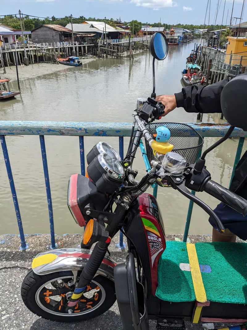Electric scooter parked on a bridge with a small rubber duck wearing a helmet attached to the handlebar, overlooking a river and stilt houses.