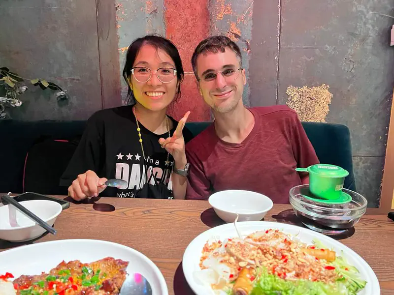 Two people smiling at a restaurant table with plates of Vietnamese food, including spring rolls and grilled meat.