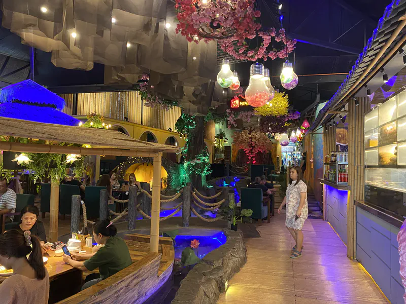 Interior of a themed restaurant with vibrant lighting, artificial cherry blossoms, colorful hanging lanterns, and customers dining near a water feature.