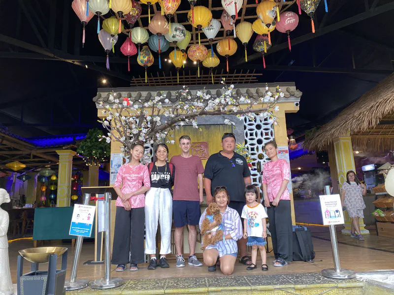 Group photo of visitors and staff standing in front of Mini Nguyen Café, with colorful lanterns hanging above and a small dog being held in the center.