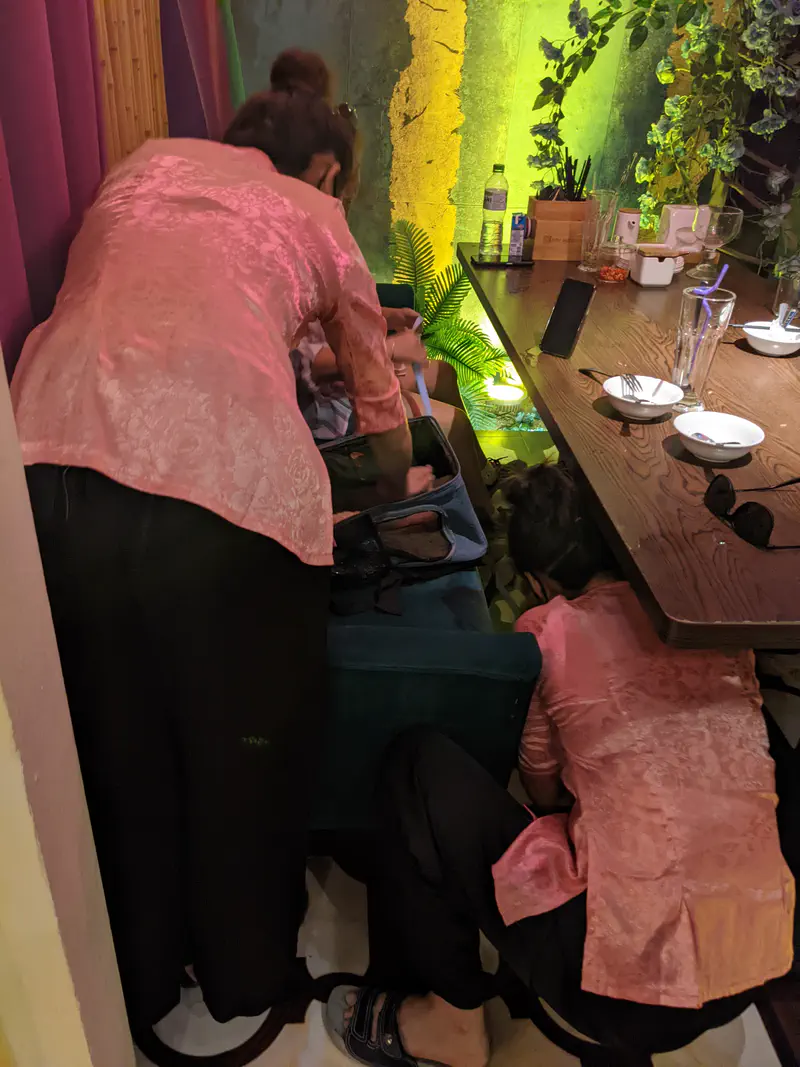 Restaurant staff in pink uniforms kneeling and helping near a dining booth while guests sit at the table.