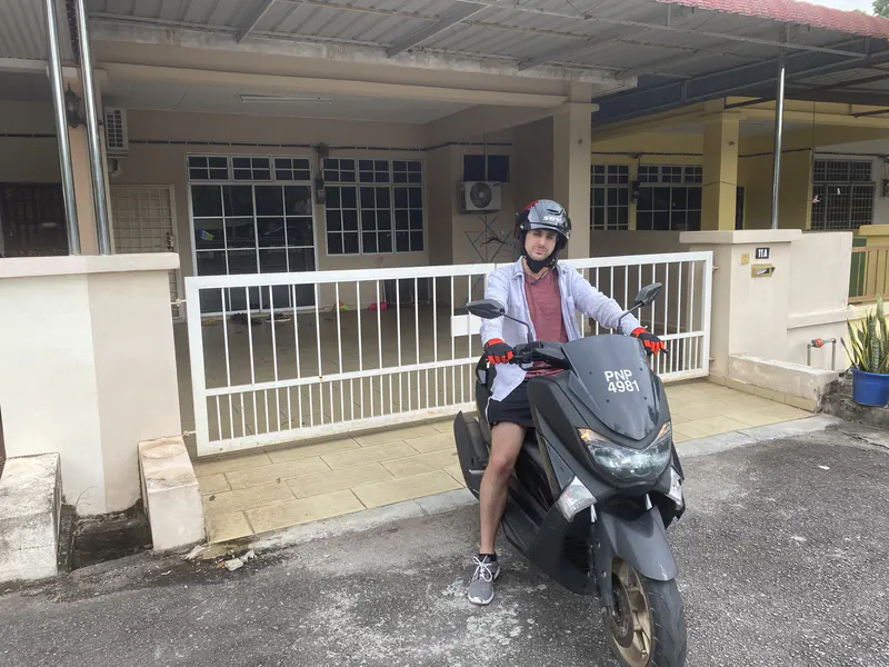 Person wearing a helmet and gloves sitting on a black scooter parked in front of a house with a gated driveway.