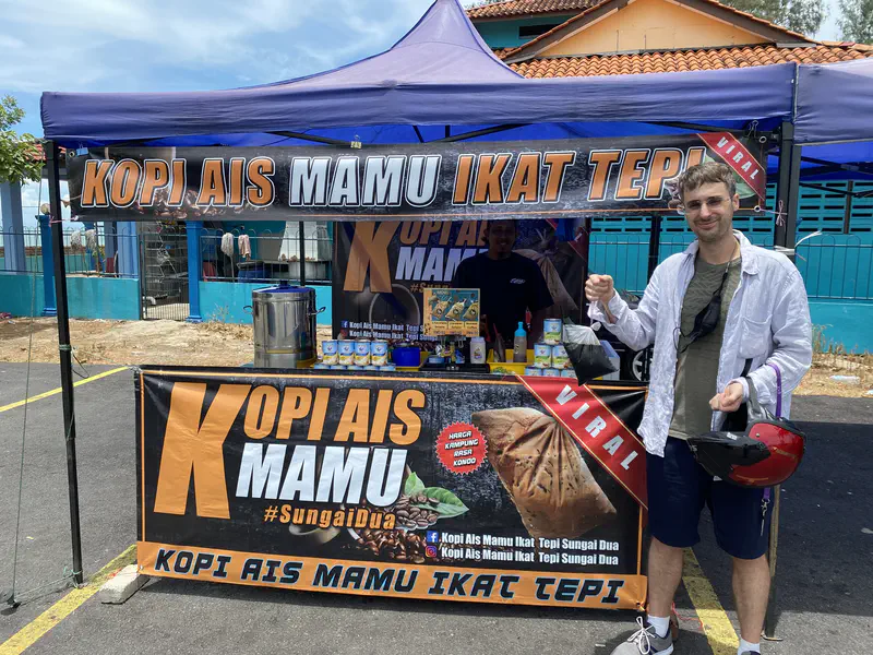 Man standing in front of a street stall called 'Kopi Ais Mamu Ikat Tepi' holding a bag of iced coffee.