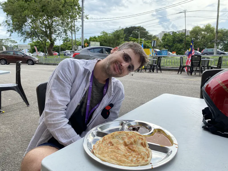 Person sitting at an outdoor table with a plate of roti canai and dipping sauces at a mamak stall.