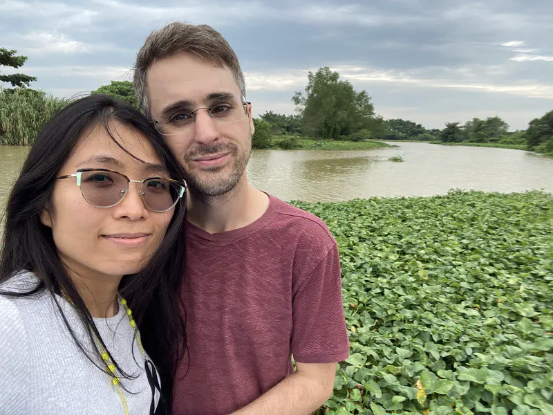 Two people smiling by a riverbank covered with green plants.