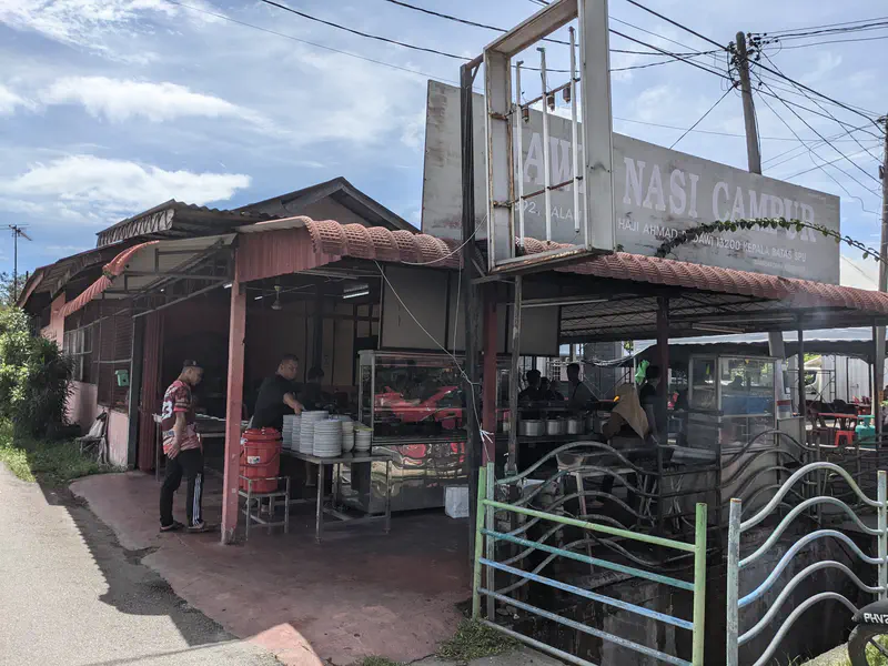 Exterior of a roadside restaurant with a sign reading 'Nasi Campur' and people preparing food at the entrance.