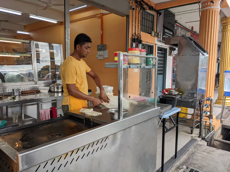 Man preparing roti canai dough at a street-side mamak stall with a hot griddle in front of him.