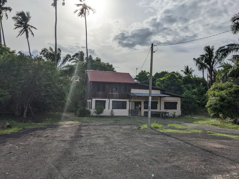 A rustic two-story house with a red roof, surrounded by tall palm trees and dense greenery, under a cloudy afternoon sky with sunlight breaking through.