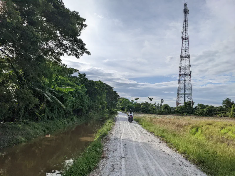 A narrow dirt road beside a canal and rice fields, with a motorcyclist riding toward a tall telecommunications tower in the background.