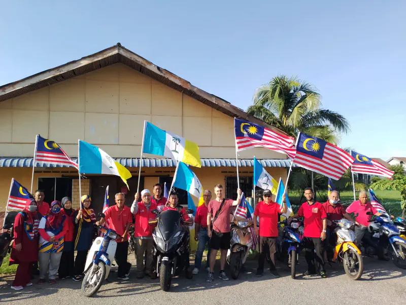 Group of people in red shirts standing with motorcycles, holding Malaysian and state flags in front of a building.